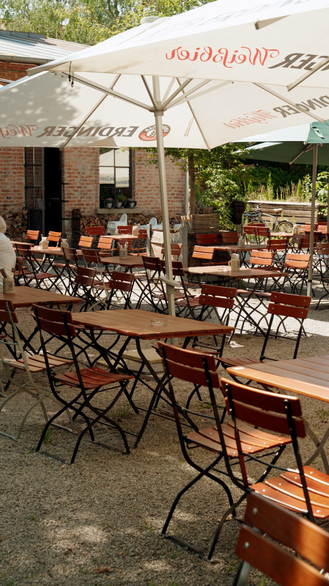 Biergarten mit weißen Schirmen, Holztischen und Bänken vor der Backstein-Fassade